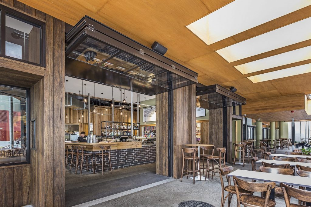 An open bar restaurant, tables, and chairs for seating with custom vertical folding doors open overhead. Connecting indoors with the outdoors beautifully. A Mixture of raw industrial and woodgrain textures throughout.
