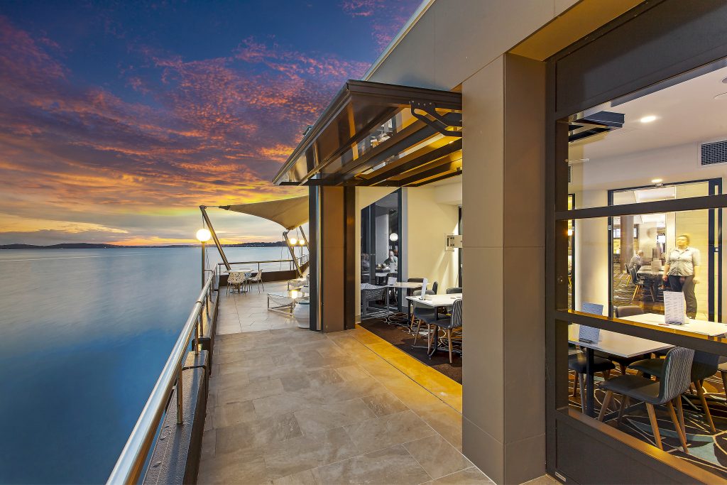 sunset, a sky filled with pink, orange and yellow clouds. a calm ocean and a tilt wall open to allow paterons to watch the sunset.